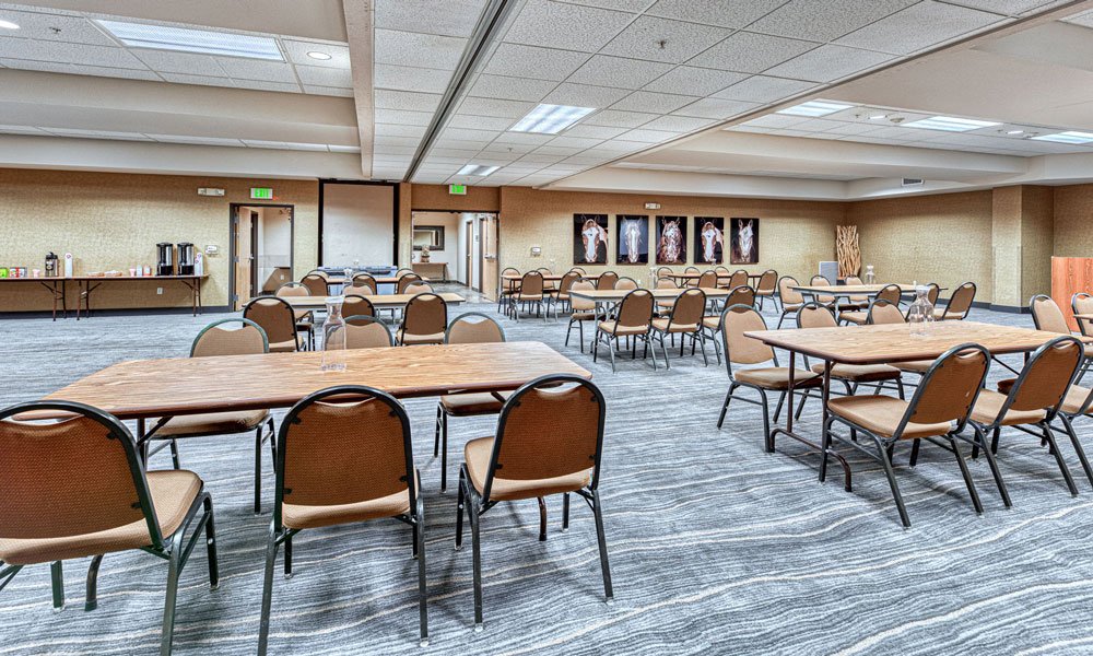 A large conference room at a hotel in Madras, Oregon, features several rectangular tables and brown chairs arranged in rows. There is a refreshment area in the corner and framed pictures on the far wall. The carpet is gray with a wavy pattern.