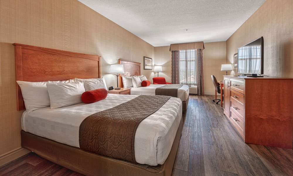 Enjoy a comfortable stay at this Hotel in Madras, Oregon, featuring a hotel room with two double beds, brown headboards, white bedding, red accent pillows, a TV on a wooden dresser, a desk chair, lamps, and a window with curtains letting in natural light.